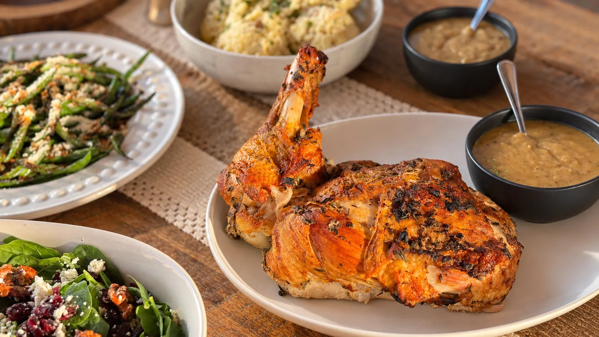 Whole roast chicken with a bowl of brown gravy, cranberry sauce, mashed potatoes, stuffing, roasted vegetables, and a green salad on a wooden table